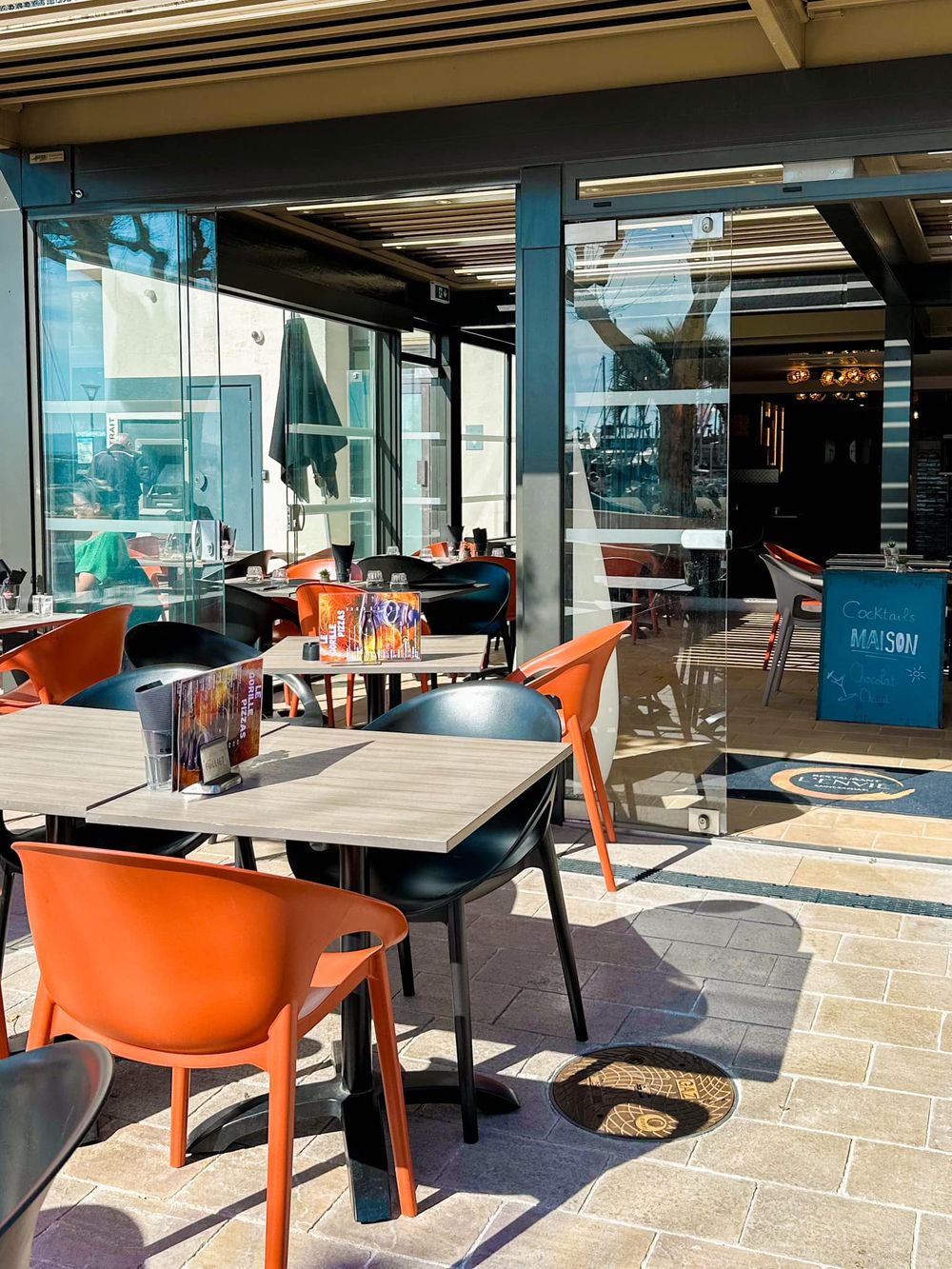 La terrasse vitrée avec ses tables en bois et ses fauteuils orange, de jour